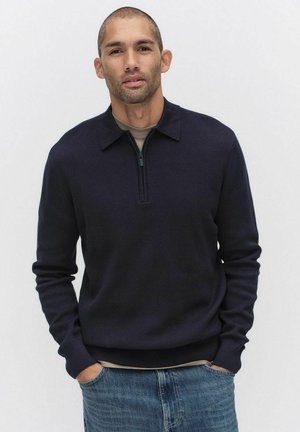 Donkerblauwe gebreide pullover met een ritskraag, geribde manchetten en zoom. Gedragen over een lichtgekleurde shirt, gecombineerd met blauwe spijkerbroek.