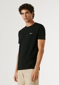 Junger Mann mit lockigem dunkelbraunem Haar, der ein schwarzes Pepe Jeans T-Shirt und beige Hosen trägt und vor einem einfarbigen hellen Hintergrund steht.