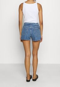 Siniset farkkushortsit, korkeavyötäröiset ja viiden taskun mallilla. Korostettu saumoilla, repeytyneillä reunoilla ja kuluneella tekstuurilla. Yhdistetty valkoiseen hihattomaan toppiin.