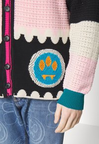 Gebreid vest met roze, zwarte en crèmekleurige panelen, voorzien van een blauwe ronde patch met een oranje pootafdruk en geribbelde groene manchetten.