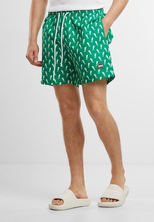 Bañadores verdes con un patrón de caballitos de mar blancos, cinturilla elástica con cordón y un pequeño parche de logo. Llevados con sandalias de color claro.