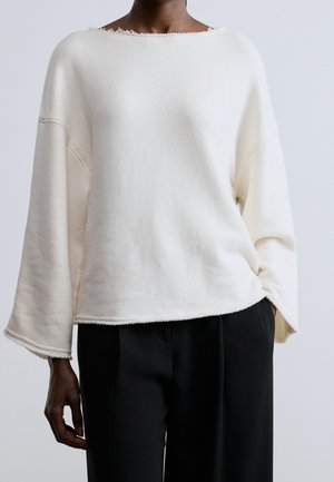 Person trägt ein lockeres weißes Langarmshirt mit rohen Kanten und schwarze Hose, steht vor einem einfachen hellen Hintergrund.