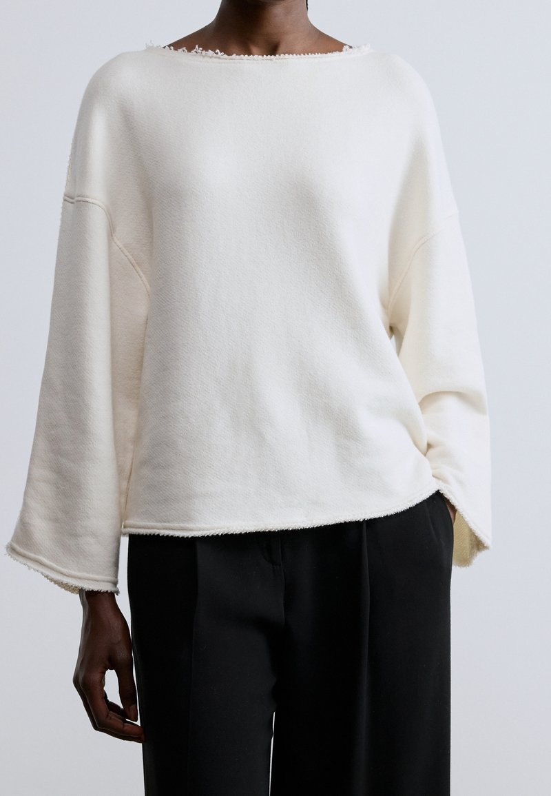 Personne portant un haut blanc à manches longues ample avec des bords bruts et un pantalon noir, debout devant un fond clair uni.