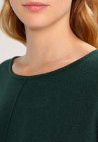 Ciemnozielony dziany sweter z okrągłym dekoltem i gładką fakturą. Szczegóły obejmują subtelny wzór szwów pod dekoltem.