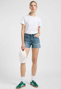 Biała bawełniana koszulka, niebieskie dżinsowe szorty z postrzępionym dołem, zielone zamszowe sneakersy z białymi paskami i biała czapka trzymana w lewej ręce.