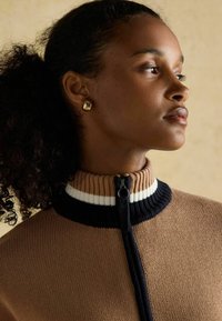 Pruunist kudumist särgist kõrge kaelusega, sinise ja valge triibuga. Tõmblukuga esine ja soonikkoes krae. Kuldsed kõrvarõngad.