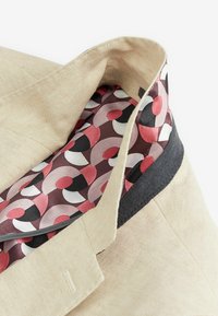 Blazer en lin beige clair avec des accents gris foncé. La doublure intérieure présente un motif géométrique dans des tons de rose, rouge et noir.