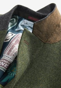 Veste en mélange de laine avec un extérieur vert, un col anthracite et une doublure bleu à motifs de tickets. Détails de couture visibles.