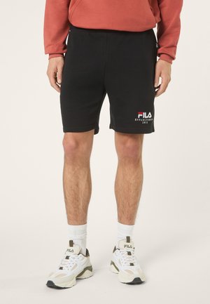 Mężczyzna ubrany w czarne spodenki FILA, białe buty FILA, białe skarpetki i czerwoną bluzę z długim rękawem, stojący na jednolitym tle.