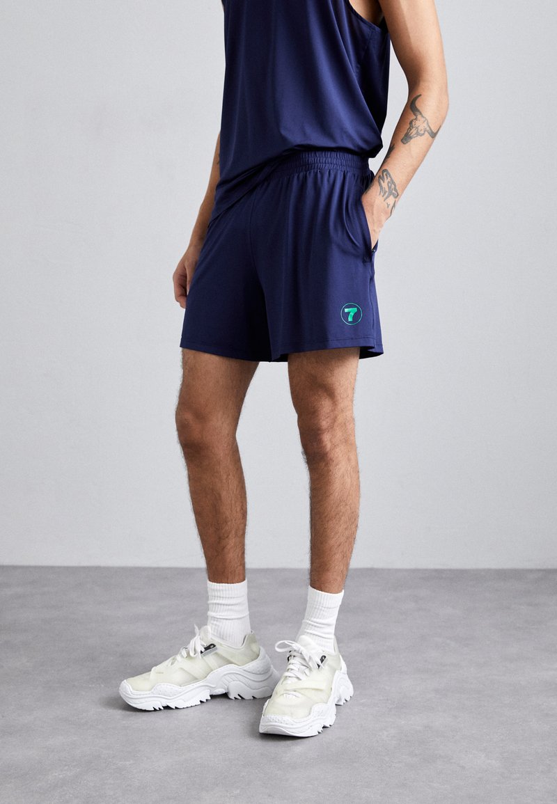 Marine treningsshorts med elastisk midje, med en liten grønn logo. Kombinert med lysfargede joggesko og hvite sokker.