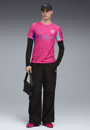 Vaaleanpunainen lyhythihainen paita, jossa on sinisiä yksityiskohtia ja graafinen muotoilu, yhdistettynä mustiin leveisiin housuihin ja vaaleanpunaisiin kengiksiin. Kannan mustaa olkalaukkua.