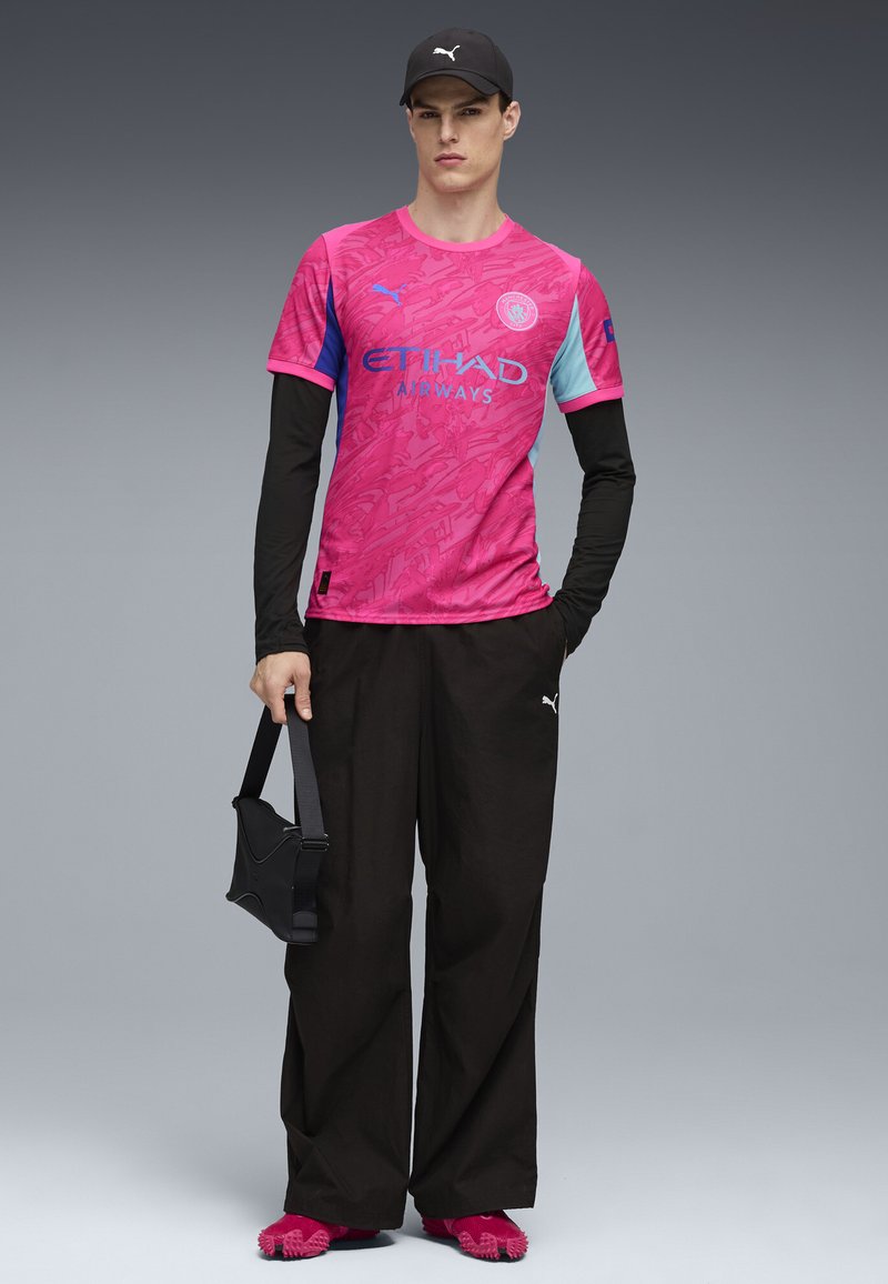 Rosa kortärmad skjorta med blå detaljer och grafisk design, parat med svarta vida byxor och rosa skor. Bär en svart axelväska.