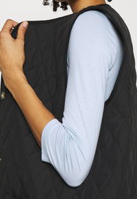 Persona che indossa una camicia a maniche lunghe blu chiaro sotto un gilet trapuntato nero, mentre aggiusta il gilet vicino alla spalla con una mano.