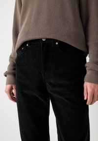 Black corduroy pants with a ribbed texture, featuring metal hardware, pockets, and a slightly tapered fit, paired with a ribbed brown sweater.