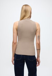 Femme aux longs cheveux blonds portant un haut beige sans manches et un jean foncé, vue de dos avec des tatouages visibles sur le bras gauche.