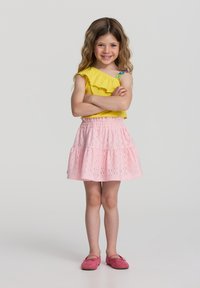 Top giallo a spalla singola con dettagli a ruches, abbinato a una gonna rosa in pizzo e scarpe rosa. Capelli lisci, braccia incrociate in una posa in piedi.