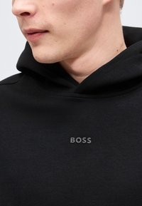 Κοντινό πλάνο ενός ατόμου που φοράει μαύρο φούτερ με κουκούλα, με το ανάγλυφο λογότυπο "BOSS" στο στήθος.