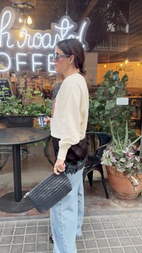 Krémový sveter s grafickým detailom, čierna sukňa s čipkovým okrajom, svetlomodré široké džínsy a čierna pletená kabelka. Okuliare a šálka kávy v ruke.