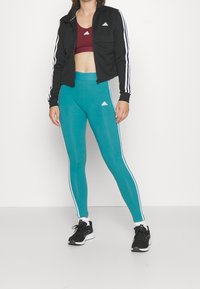 Svart zip-up jacka med vita ränder, vinröd sport-bh och teal leggings med vita sidosträck, tillsammans med svarta träningsskor.