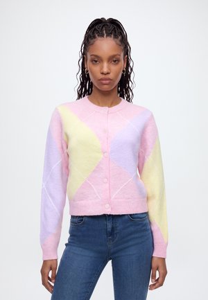 Kvinde iført en pastelfarvet cardigan med pink, gul og lavendel geometriske mønstre kombineret med blå jeans, stående mod en hvid baggrund.