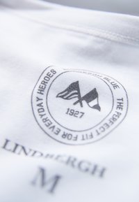 Weißer Stoff mit einem kreisförmigen Logo, das zwei Fahnen zeigt, den Text "EVERYDAY HEROES", "1927" und den Markennamen "LINDPERGH M" in Grau.