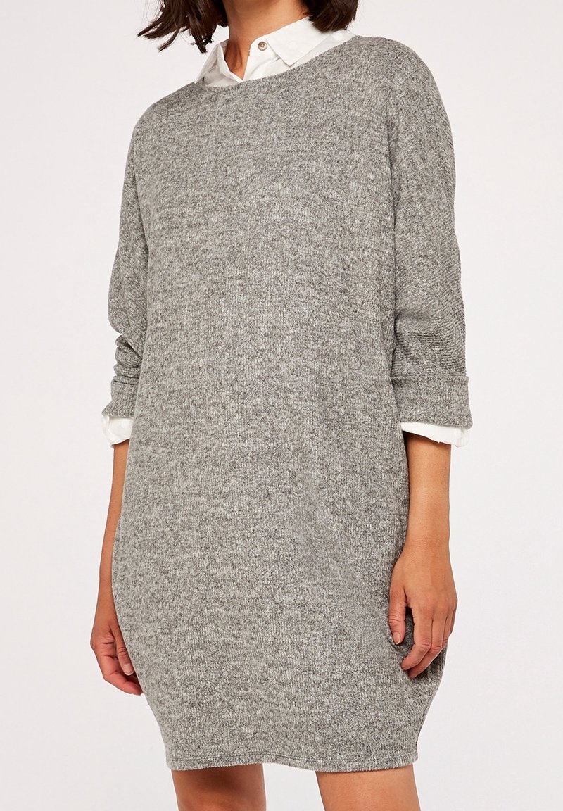 Femme portant une robe-pull grise à manches longues en tricot par-dessus une chemise blanche à col avec les manches retroussées.