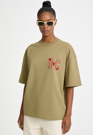 Frau trägt ein hellbraunes übergroßes T-Shirt mit rotem Herz und X-Tic-Tac-Toe-Design, cremefarbene Hose und hellfarbene rechteckige Sonnenbrille.