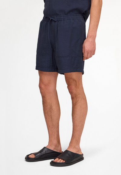 Male wearing navy elastic waist shorts and black slip-on sandals, standing against a plain white background.