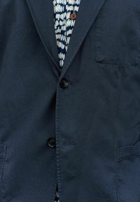 Blazer blu navy con due bottoni, rever a toppa e tasca sul petto. Tessuto a trama, abbinato a una camicia a righe blu e bianca sotto.