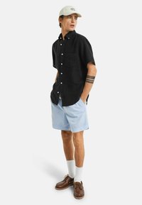 Schwarz kurzärmeliges Hemd mit Knöpfen, hellblaue Shorts, weiße Socken und braune Schuhe. Die Mütze ist hellbeige mit Logodetail. Ein Tattoo ist am Arm sichtbar.