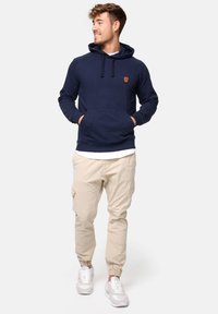 Sudadera azul marino con bolsillo tipo canguro, capucha con cordones y parche de logo marrón, combinada con pantalones cargo beige y zapatillas blancas.