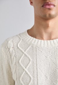 Cremfarbener Strickpullover mit einem Kabeldesign auf der Vorderseite und einem gerippten Halsausschnitt. Strukturierter Stoff mit einem gemütlichen Erscheinungsbild.