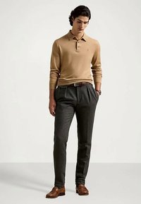 Man med långärmad polotröja i kamelfärg, mörkgrå plisserade byxor, brun bälte, bruna läder loafers, står med händerna i fickorna mot en neutral bakgrund.