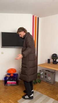 Casaco puffer castanho longo com colarinho alto e costuras horizontais. O interior é suave e atinge o comprimento até metade da panturrilha. Calças pretas e sandálias.