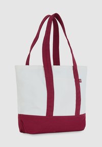 Biała torba tote z burgundowymi ramiączkami i dnem, stojąca na neutralnym tle.