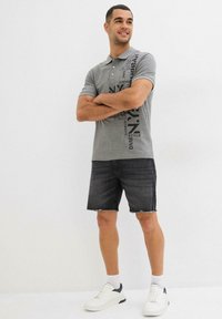Polo grigio con testo grafico nero, abbinato a pantaloni shorts in denim neri. Sneakers bianche con dettagli blu navy completano l'outfit.