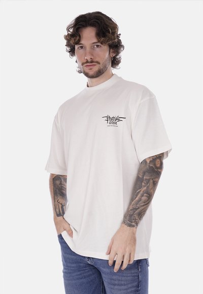 T-shirt bianca in cotone con vestibilità rilassata, maniche corte e una piccola grafica nera sul lato sinistro del petto. Indossata con jeans blu.