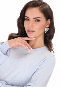 Maglione azzurro chiaro con maniche lunghe, scollo tondo e dettagli con bottoni sulla spalla. La modella ha capelli castani lunghi e indossa orecchini di perle.