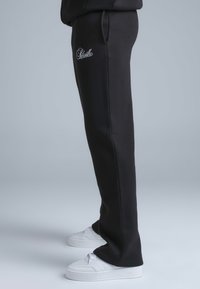 Pantalon de survêtement noir à coupe slim, avec un logo blanc brodé et des poches latérales, associé à des baskets blanches sur un fond gris.