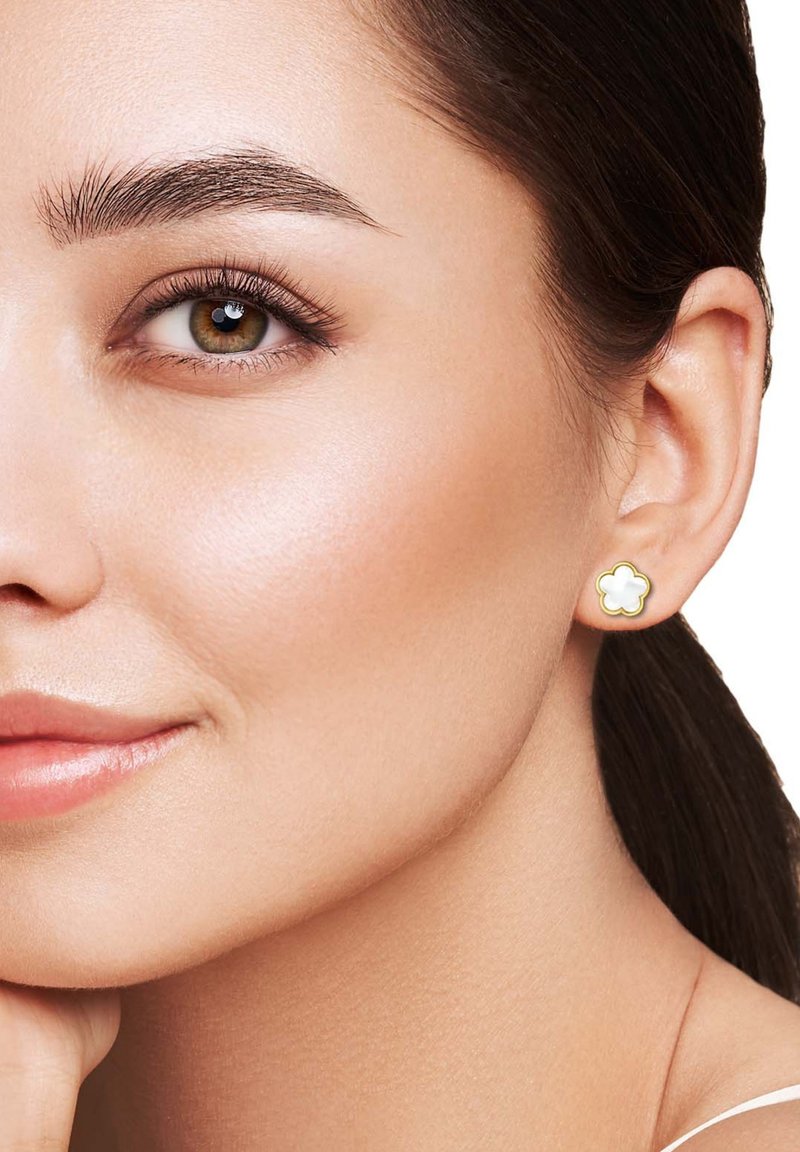 Boucle d'oreille en forme de fleur ton doré avec un centre en émail blanc, présentant un bord festonné et une finition polie, portée à l'oreille d'un modèle.