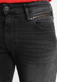 Pantalones vaqueros de mezclilla negros con un bolsillo con cremallera, diseño clásico de cinco bolsillos y un sutil desgaste en la textura del tejido.