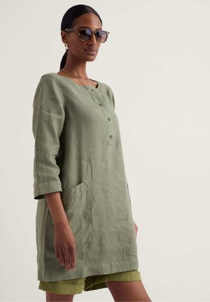 Kobieta w oliwkowo-zielonej lnianej tunice z guzikami z przodu, kieszeniami i rękawami 3/4, w połączeniu z pasującymi szortami i okularami przeciwsłonecznymi.