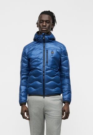 Mees, kes kannab säravat sinist kapuutsiga tikitud jopet, millel on ees tõmblukk, ja helehallid püksid, seisab tavalise tausta ees.