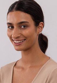 Nahaufnahme eines Porträts einer Person mit mittlerem braunen Hautton, die ein geripptes beigefarbenes Oberteil und kleine Perlenohrringe trägt, mit in einem tiefen Pferdeschwanz gestyltem Haar.