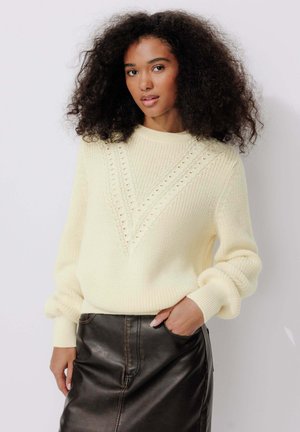 Pull en crème texturé avec un motif en V et des manches amples, associé à une jupe en cuir noir.