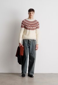 Gestrickter Pullover mit geometrischem roten und cremefarbenem Muster, kombiniert mit grauen, weit geschnittenen Jeans und schwarzen Schuhen. Das Model hält einen braunen Pullover.