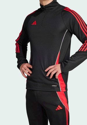 Osoba nosząca czarno-czerwony strój sportowy Adidas z długimi rękawami, kołnierzem na zamek, trzema czerwonymi paskami na rękawach i spodniach oraz logo Aeroready.