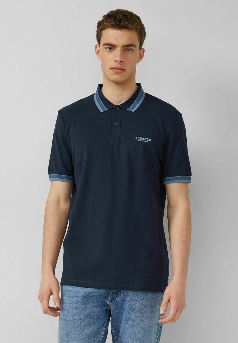 Junger Mann trägt ein marineblaues Poloshirt mit hellblauem Kragen und Ärmelsaum, kombiniert mit hellblauen Jeans, steht vor einem grauen Hintergrund.