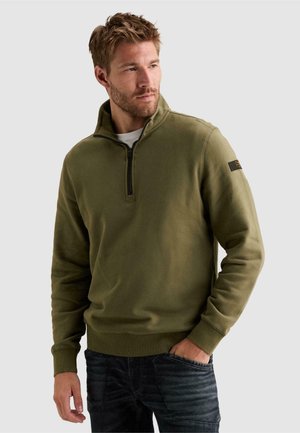 Homme portant un pull vert olive à fermeture éclair quart de zip avec un écusson sur la manche, regardant sur le côté avec une main dans la poche.