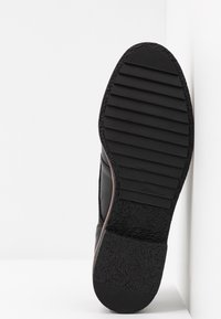 Semelle de chaussure noire avec des rainures horizontales à l'avant et un talon texturé, photographiée sur un fond blanc uni.
